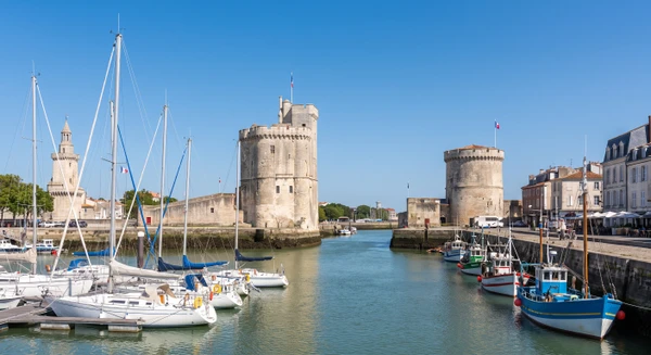 Vue panoramique de la façade maritime de La Rochelle avec ses trois tours historiques (Tours de la Chaîne, de la Lanterne et de la Grosse Horloge) visibles depuis le port, avec des bateaux en premier plan et le ciel dégagé. Image claire et reconnaissable, de qualité professionnelle, représentative de la ville sans aucun texte, logo ou filigrane.